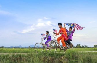 Boys in traditional clothes riding bikes waving Malaysian flags in green rural field