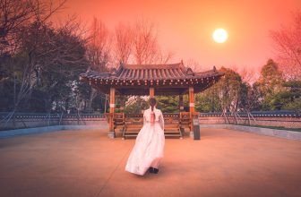 Woman in hanbok walking toward traditional Korean pavilion at sunset