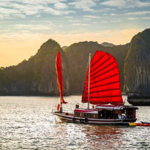 Traditional Vietnamese junk boat with red sails near limestone cliffs at sunset