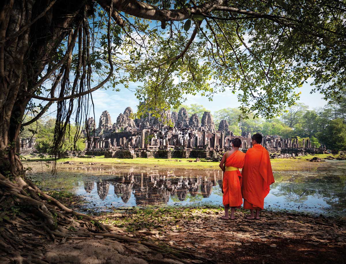 Two monks in orange robes standing beside a pond reflecting ancient temple ruins under trees