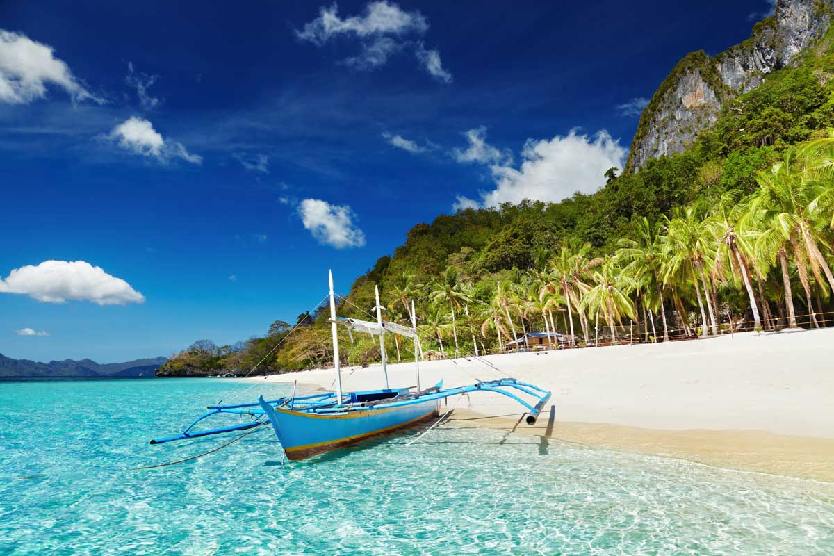 Blue outrigger boat on turquoise water at tropical beach with palm trees and rocky hill