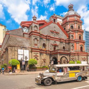 Historic stone church with silver jeepney on city street under blue sky