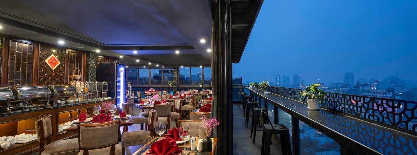 Rooftop restaurant and bar with tables and chairs overlooking Hanoi city at dusk.