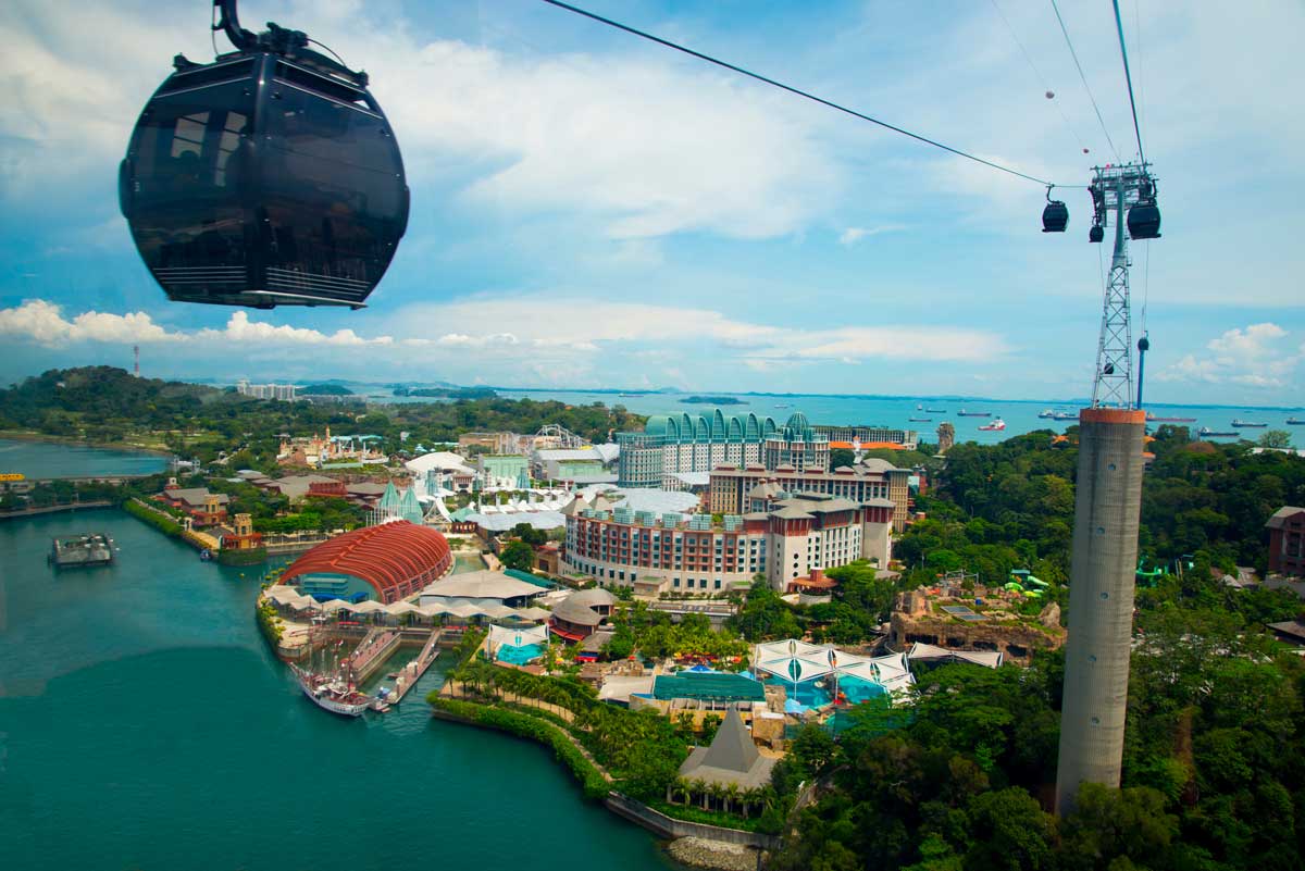 Cable cars over resort and water with buildings and greenery under partly cloudy sky