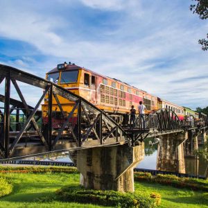 Colorful train crossing iron railway bridge over river with passengers and greenery