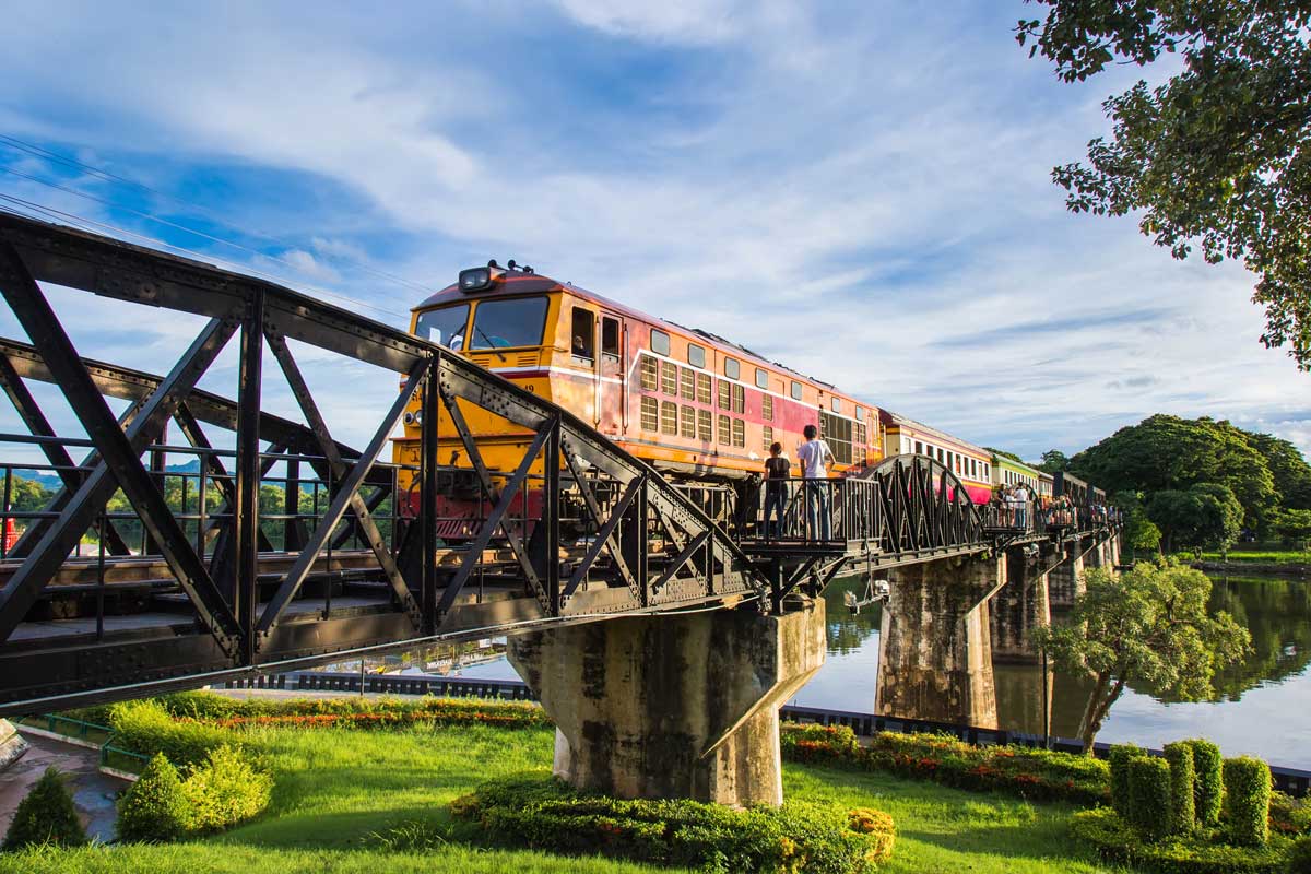 Colorful train crossing iron railway bridge over river with passengers and greenery