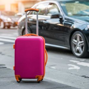 Pink suitcase on city sidewalk next to black car and traffic