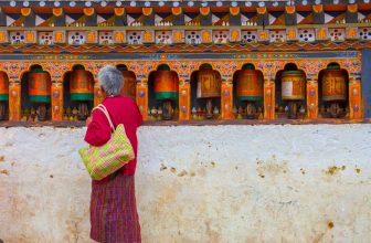 Woman in traditional dress standing by colorful Bhutanese prayer wheels on decorated wall