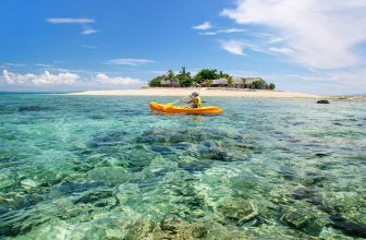 Person kayaking on clear water near a tropical sandy island with palm trees