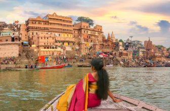 Woman in pink and yellow sari seated on boat looking at the Ganges River ghats in Varanasi