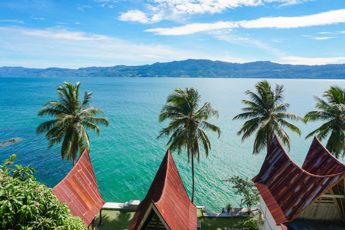 Red triangular roofs and palm trees overlooking a large blue lake with mountains beyond under blue sky