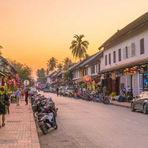 Street at sunset with motorbikes, shops, palm trees, and people walking