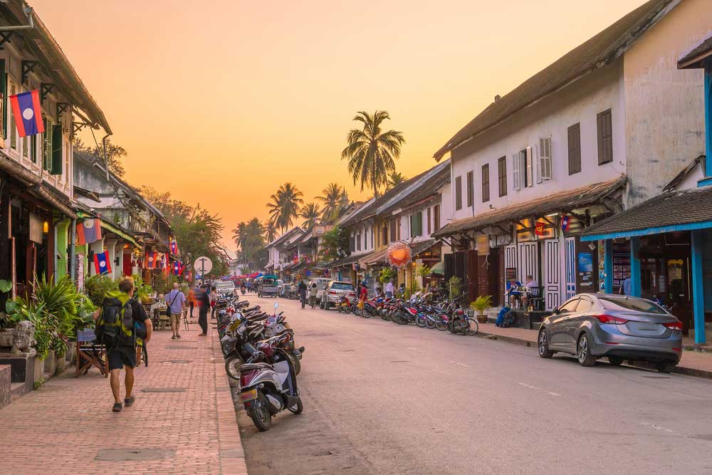 Street at sunset with motorbikes, shops, palm trees, and people walking