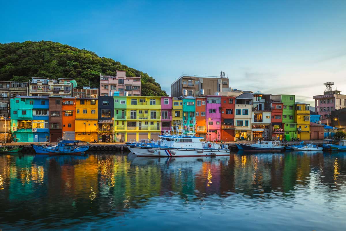 Colorful buildings by harbor with boats and green hillside in background under clear sky