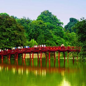 People walking on a red wooden bridge over a green lake surrounded by lush trees