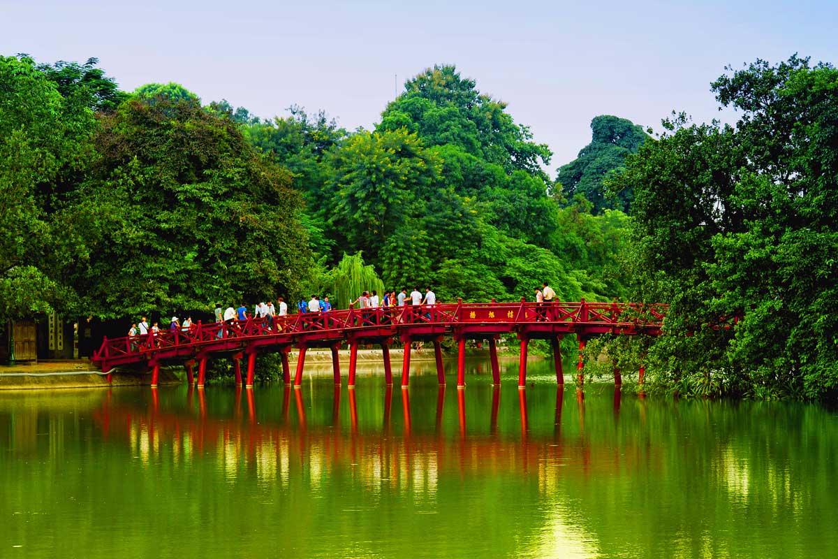 People walking on a red wooden bridge over a green lake surrounded by lush trees