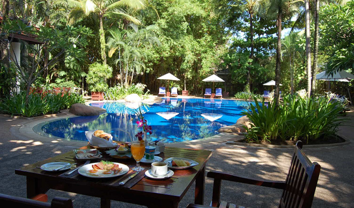 Poolside breakfast table near curved pool surrounded by palm trees and sun loungers