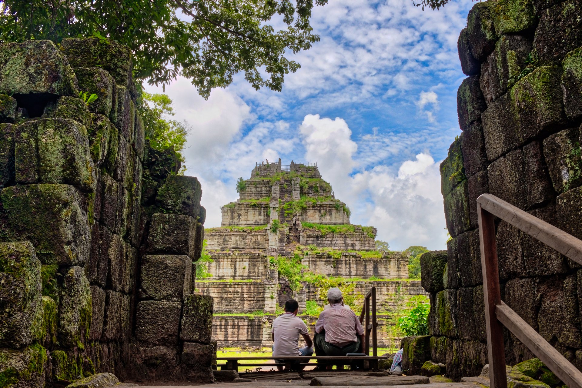 Two people sitting on bench facing moss-covered stepped pyramid temple ruins