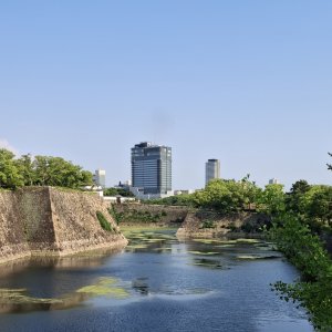 Stone walls and water moat beside green trees with tall buildings in background
