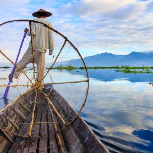 Fisherman balancing on boat using circular fishing net on calm lake water