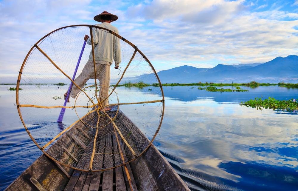Fisherman balancing on boat using circular fishing net on calm lake water