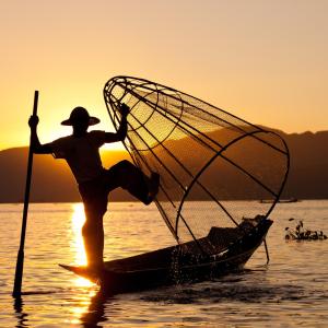 Silhouette of fisherman using conical net on boat at sunset on calm lake