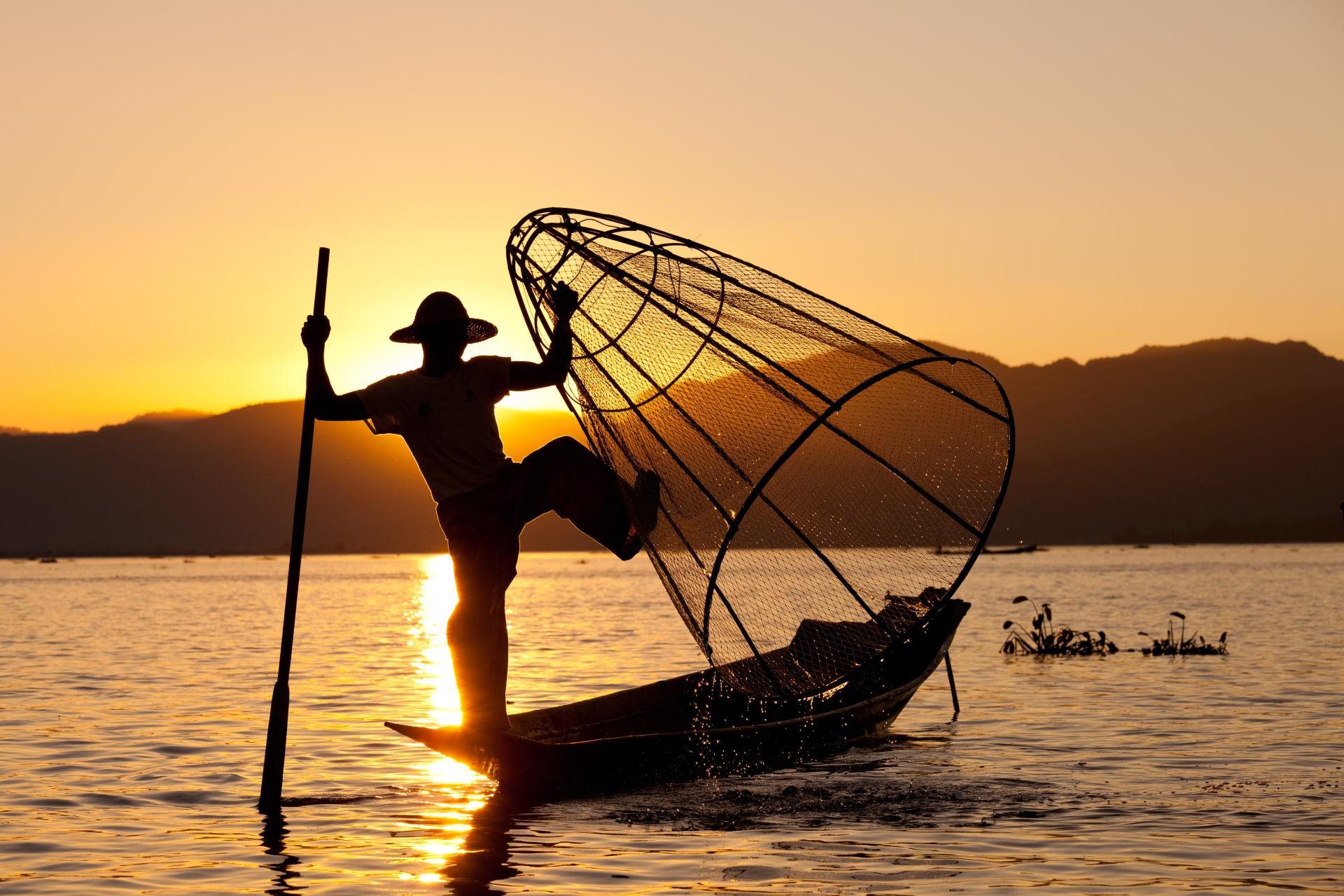 Silhouette of fisherman using conical net on boat at sunset on calm lake