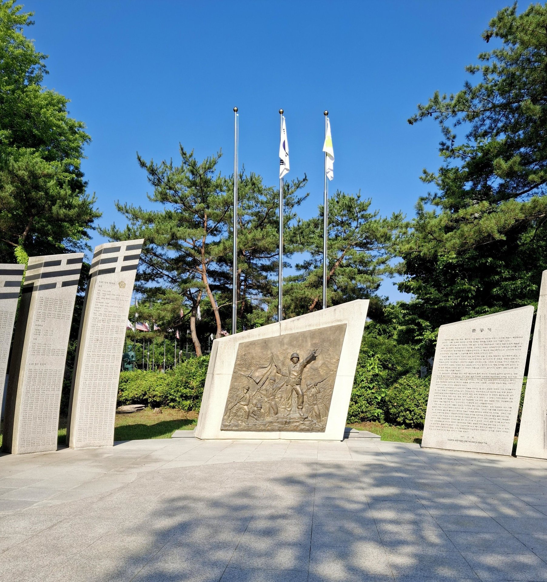 Memorial with soldier relief, stone inscriptions, flagpoles, and pine trees under clear sky