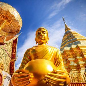 Golden Buddha statue with temple and gilded stupa under blue sky in Thailand