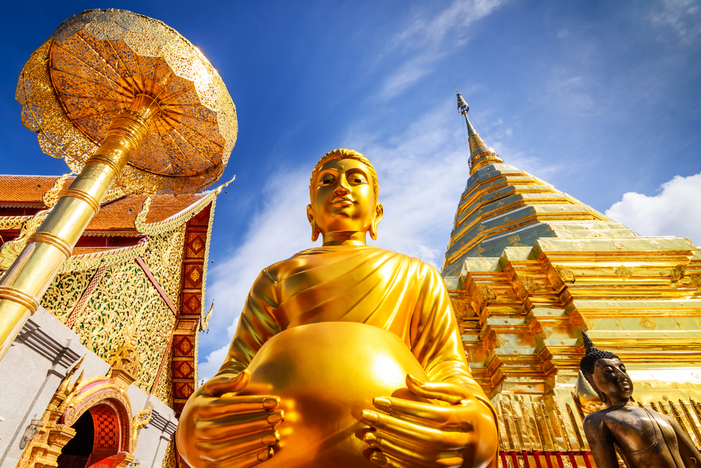 Golden Buddha statue with temple and gilded stupa under blue sky in Thailand