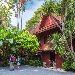 Two people with backpacks walking near a traditional red Thai wooden museum surrounded by lush plants.