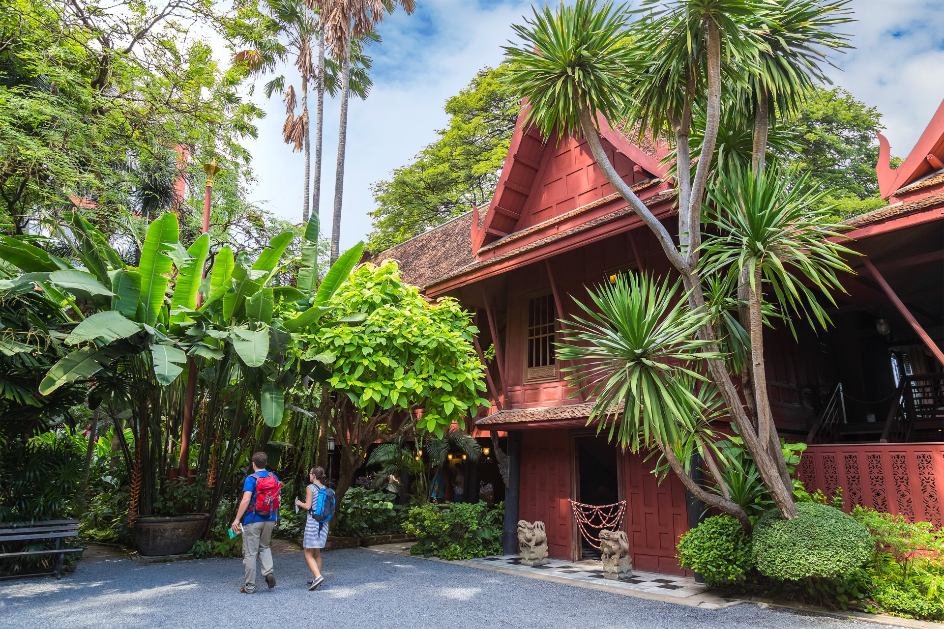 Two people with backpacks walking near a traditional red Thai wooden museum surrounded by lush plants.