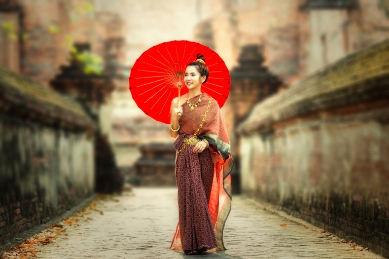 Young woman in traditional Thai dress holding red parasol in historic temple corridor