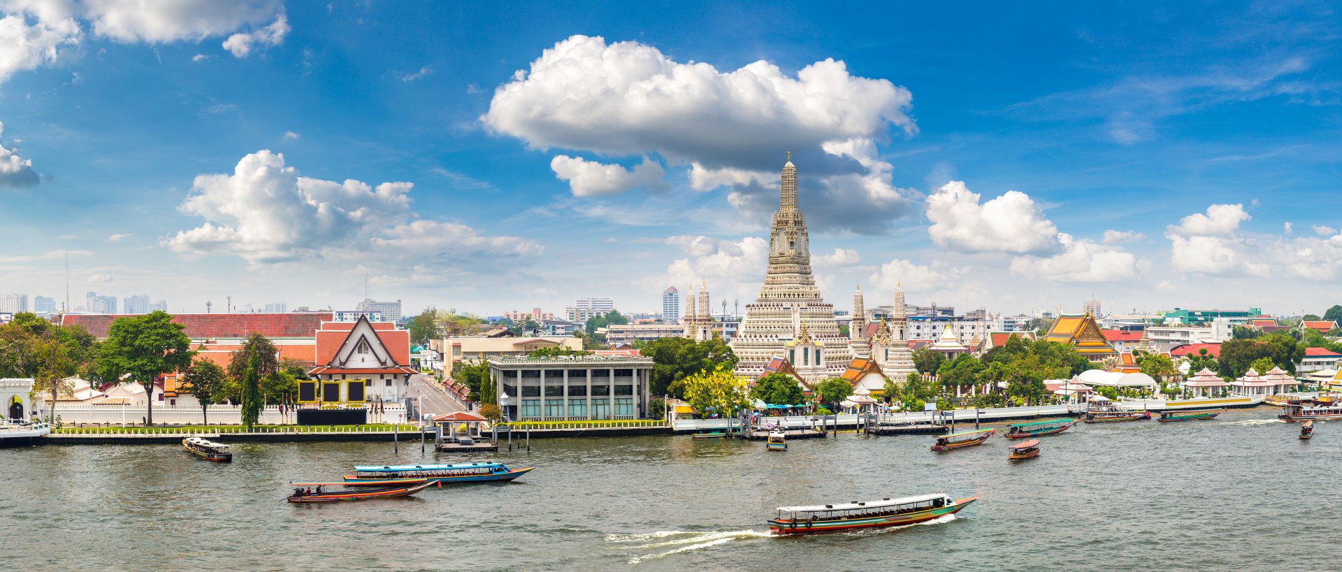 Wat Arun temple by river with boats and cityscape under blue sky