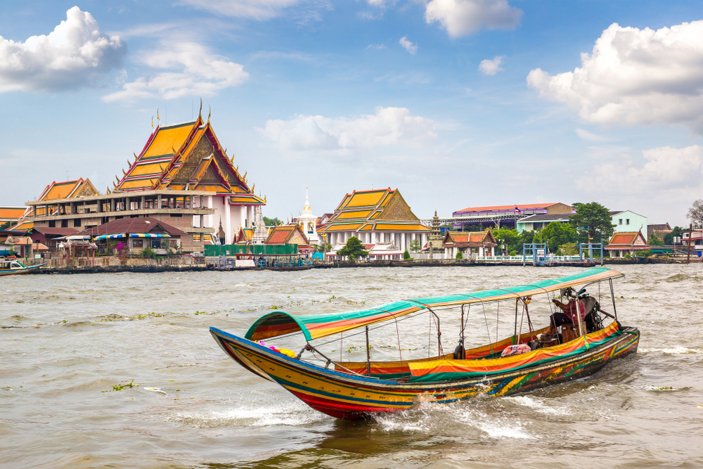 Colorful long-tail boat on river in front of traditional Thai temples with orange roofs