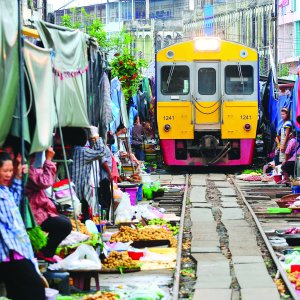 Yellow train passing through crowded open-air market with people and fresh produce on tracks