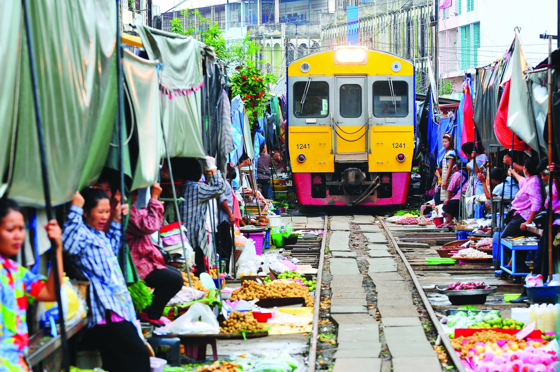 Yellow train passing through crowded open-air market with people and fresh produce on tracks