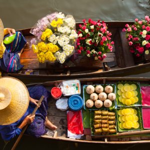 Two vendors in traditional hats sell flowers and fruits from wooden boats on water