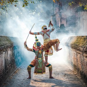 Two masked Khon dancers performing in colorful traditional costumes in a misty brick alley