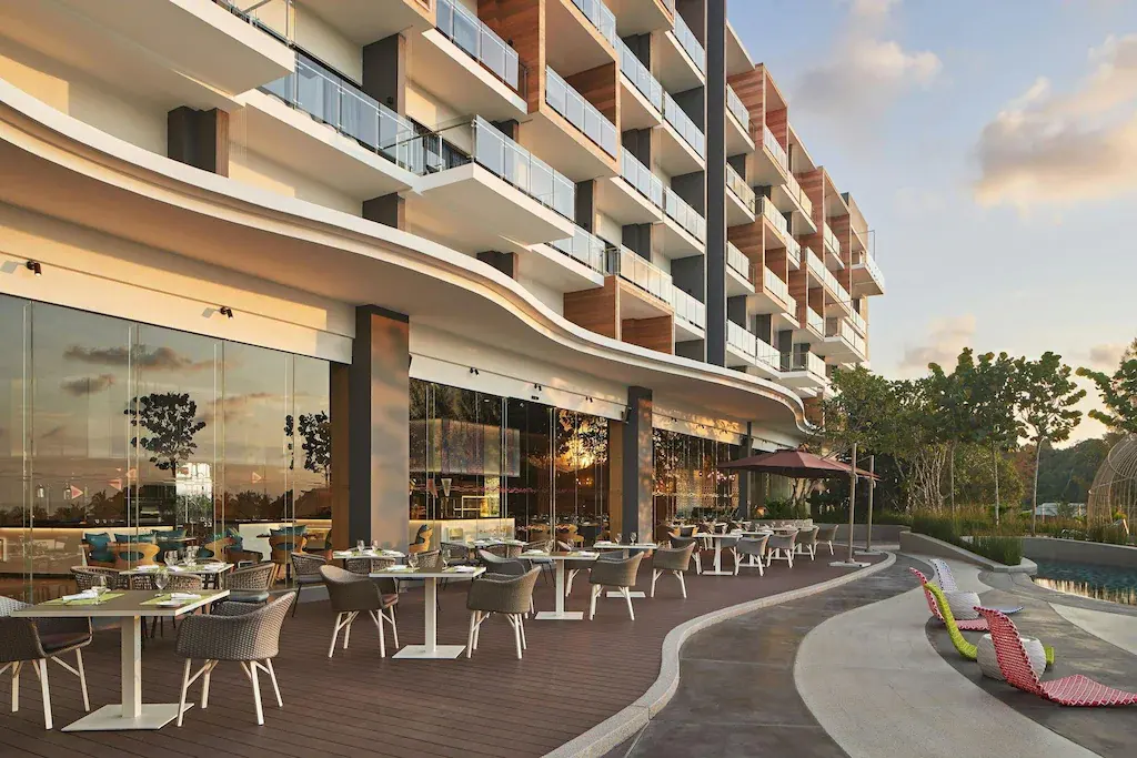 Outdoor dining area with tables and chairs beside Aloft Langkawi Pantai Tengah resort building
