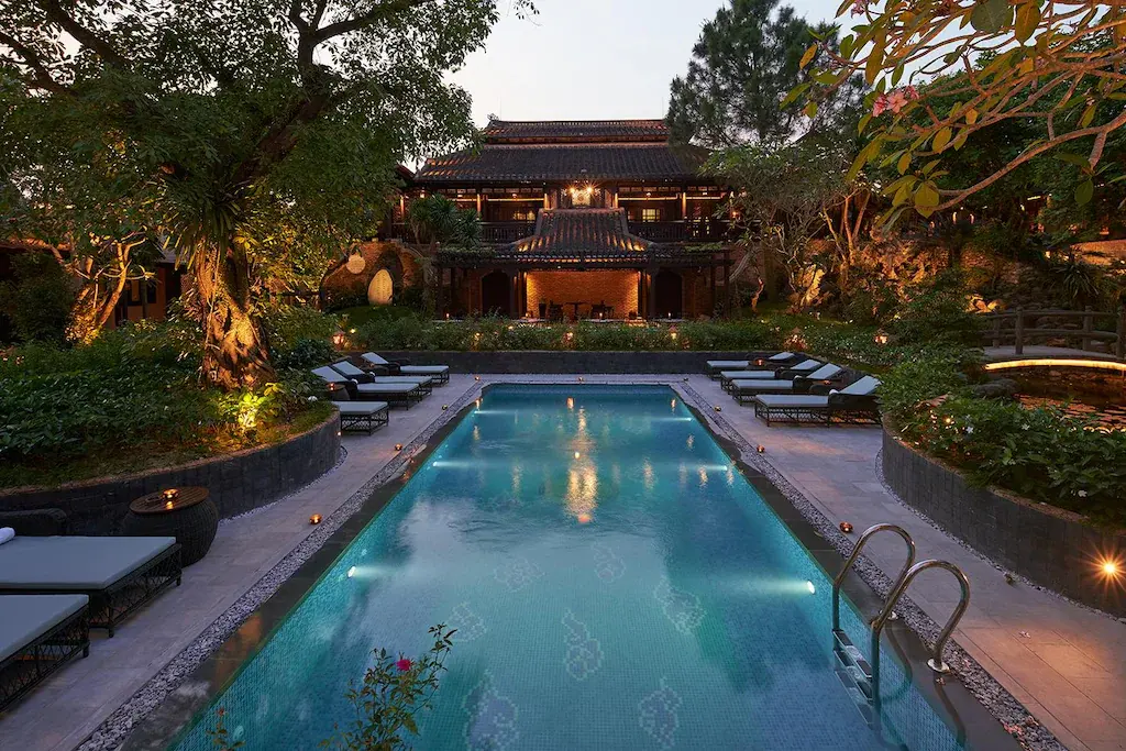 Traditional garden house with illuminated swimming pool at dusk surrounded by trees and plants