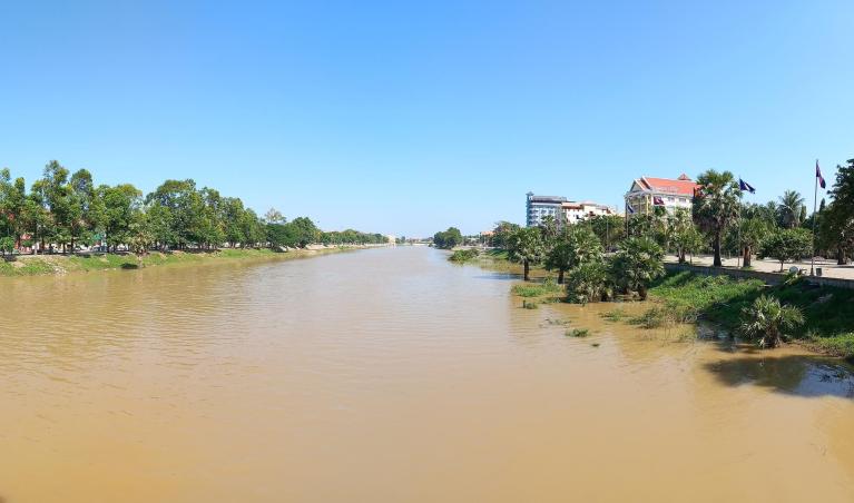 Riverside view with palm trees and buildings along muddy river under clear blue sky in Battambang, Cambodia