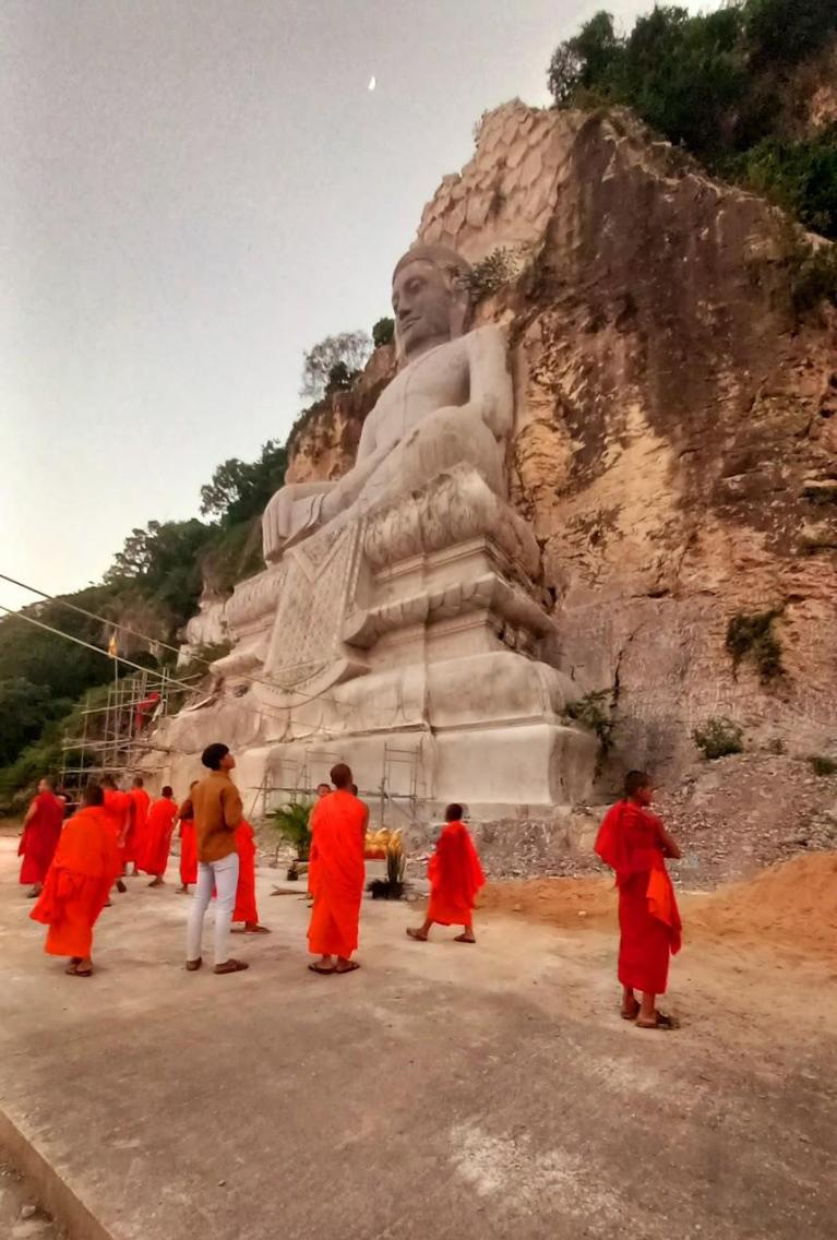 Monks in orange robes standing near a large Buddha statue carved into a rocky cliff in Battambang Cambodia.