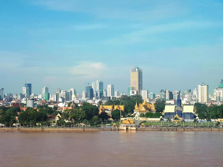 Phnom Penh city skyline with Mekong River waterfront and golden-roofed buildings under blue sky.