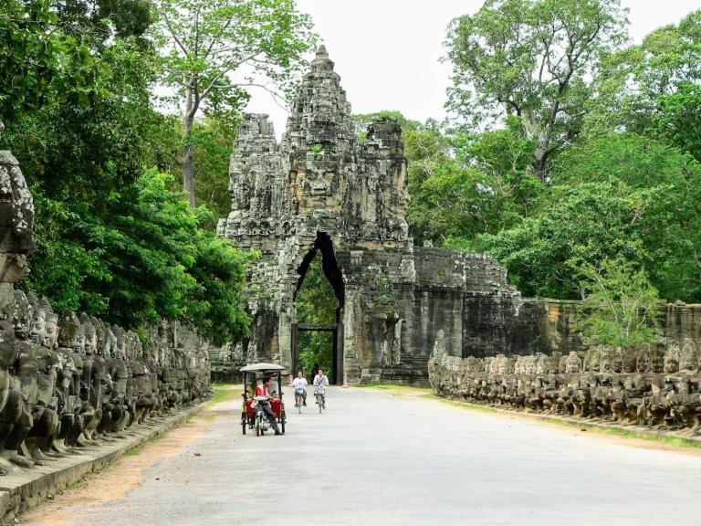 Stone gateway with tuk tuk and cyclists on road surrounded by green trees in Siem Reap