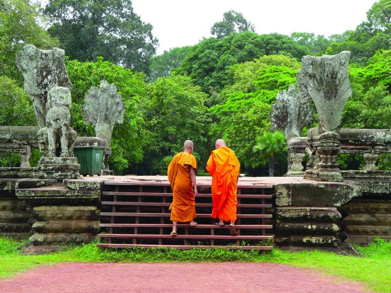 Two Buddhist monks in orange robes walking up stone steps with ancient sculptures in Siem Reap