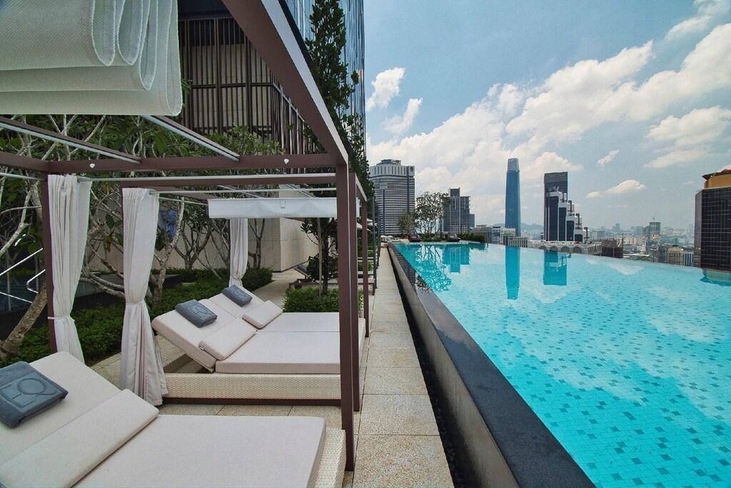 Rooftop infinity pool at EQ Kuala Lumpur with lounge chairs and city skyline under partly cloudy sky.