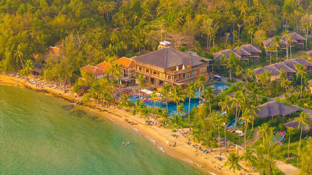 Aerial view of a tropical beachfront resort with pool, palm trees and sandy beach.