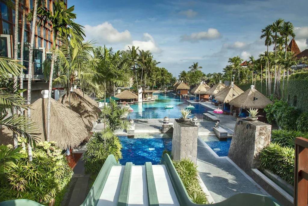 Tropical resort pool with water slides, palm trees, and thatched-roof huts