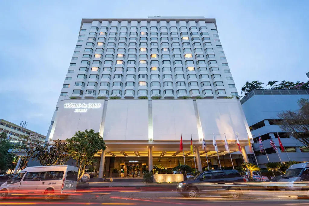 Tall hotel facade lit at dusk with cars and flags in front